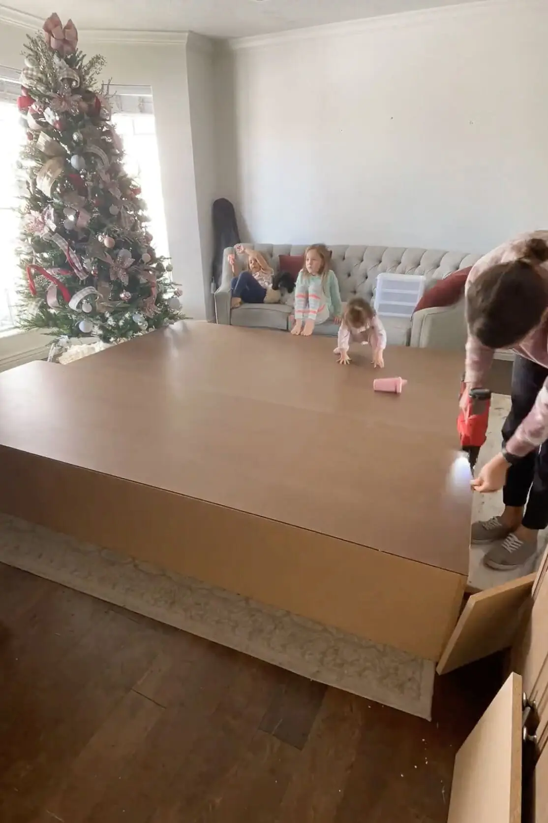 A DIYer attaches the back panel to a large storage unit while children sit nearby watching the progress in a festively decorated living room.